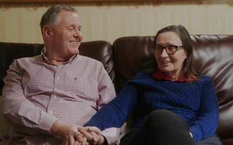 Bill and Jo sat on their couch, smiling and holding hands